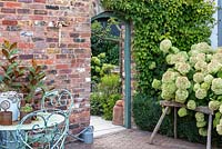The entrance to a contemporary walled cottage garden flanked by Hydrangea 'Limelight' and a seating area with vintage table and chairs.
