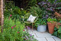 A modern chair and a terracotta pot of white violas, and galvanised metal agricultural vessel planted with pelargoniums. Behind, a raised corner bed planted with Anemone, grasses, ferns and Cercis canadensis.