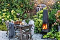 A dining area in a courtyard garden lit by a string of bulbs and candles, heated by a modern outdoor chimenea. Planting behind includes Hydrangea 'Limelight', Rosa 'Blush Noisette', Anemone hybrida 'Elfin Swan' and ferns.