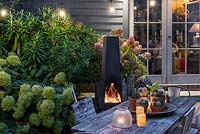 A dining area in a courtyard garden lit by a string of bulbs and candles, heated by a modern outdoor chiminea. Planting behind includes Hydrangea 'Limelight', Rosa 'Blush Noisette' and ferns.