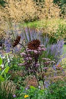 A rusty metal sculpture of a flower, set in a border amongst Stipa gigantea, Russian sage, tobacco plants and purple Angelica.