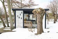 Gardenhouse with comfortable lounge furniture near frozen lake in the middle of snow. Big knotted willow tree in front