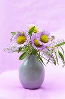 Grey vase holding Hebe and Erigeron 'Sea Breeze Mauve' flowers with a mauve background