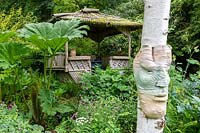 Ceramic face on tree trunk with view to summerhouse set in lush garden with bold foliage