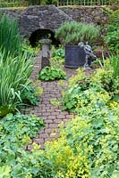 View along paved path with Alchemilla mollis to sundial, statue and container, beyond stone bridge