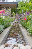Looking down a contemporary rill water feature lined with large pebbles towards modern covered outdoor room. The Viking Cruises Lagom Garden - Hampton Court Flower Festival 2019