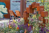 Mixed hot summer border including purple Salvia sylvestris 'Mainacht' in front of 'Honesty' sculpture in Corten steel. The Lower Barn Farm Outdoor Living Garden - Hampton Court Flower Festival 2019 