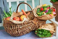 Basket with apples, carrots, spring onions, spearmint and wrapped tulips displayed on bench.
