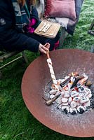 Woman wrapped in warm clothes, cooking camp fire bread over a fire in a Corten Steel fire pit on a frosty winter day