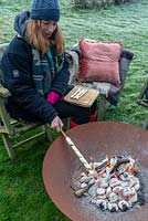 Woman wrapped in warm clothes, cooking camp fire bread over a fire in a Corten Steel fire pit on a frosty winter day