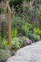 Compacted path in front of a colourful bed of summer flowering perennials with inscribed wooden post. The Cancer Research UK Pledge Pathway to Progress - Hampton Court Flower Festival, 2019.