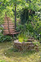 Bespoke leaf-shaped chair in wild garden with naturalistic planting and a central circular stone water feature. Calm Admidst Chaos, Hampton Court Flower Festival, 2019.