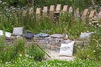 View through wildlife garden towards drystone wall seating area with cushions.  Springwatch Garden, Hampton Court Flower Show, 2019.