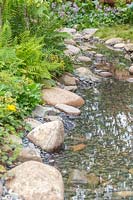 Shallow stream in woodland garden with edging of rocks and pebbles. The Zoflora and Caudwell Children's Wild Garden.  RHS Hampton Court Palace Flower Show, 2017. Designers: Adam White and Andree Davies. 