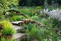 Wooden steps with insect and wildlife friendly log walls set within naturalistic borders planted with perennials and ornamental grasses.