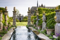 The Collector Earl's Garden at Arundel Castle, West Sussex in May. Designed by Isabel and Julian Bannermann. Green oak urns with gilded lion spouts and agaves line water representing an allegorical River Arun, leading towards Oberon's Palace. Terracotta pots are planted with agapanthus and surrounded by Alchemilla mollis and alliums.