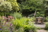 Simple dining deck surrounded by trees and soft planting including clipped, variegated standard hollies, foxgloves and yellow Sisyrinchium striatum in a cottage garden in June