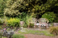 A pair of orange metal garden seats surrounded by lush planting including Prunus laurocerasus, hollies, creeping thymes, yellow flowered Lysimachia punctata and orange Carex testacaea in a cottage garden in June