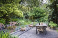 Simple decked dining area in a woodland garden surrounded by dense trees and shrubs in June. Planting includes ferns, bamboos and grasses.