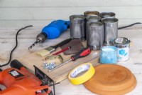 Tin cans, drill, jigsaw, wooden board, screwdriver, screws, brackets, pencil, ruler, paintbrush, paint, sandpaper, masking tape and terra cotta dish laid out on a wooden surface