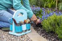 Pastel tin can caddy on the ground containing gardening tools and supplies with woman gardening in the background