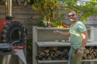 Man making pizza in outdoor kitchen with red pizza oven and built-in wood storage 