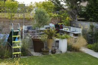 Play area with colourful plastic slide and paddling pool next to outdoor kitchen surrounded by plants in big pots and pleached trees for privacy in modern family garden 