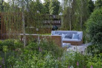 The Viking Friluftsliv Garden. Glimpse between birch tree and osmanthus to a seating area and outdoor kitchen beyond.