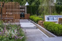 The Viking Friluftsliv Garden. A path and box-edged rill separate an outdoor dining and kitchen from sofas in a seating area shaded by birch trees.