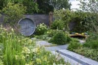 A Place to Meet Again. Hampton Court Flower Festival 2021. An urban courtyard is planted in restful tones of grey, green and white, with height added by a pergola draped in star jasmine. A circular water feature on the wall upcycles pipes and taps.