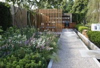 View of The Viking Friluftsliv Garden where a long path leads to an oudoor kitchen and pergola, it is bordered on the left with mixed planting including Pinus mugo 'Mops', and on the right, a long and narrow rill - Designer: Will Williams -Sponsor: Viking