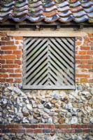 Detail of decorative shutter on window in weathered brick and flint outbuilding.
