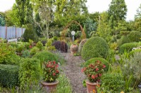 Informal garden with Fuchsia 'Thalia' in pots, clipped topiary mixed, view along gravel path to urn framed by rusty arch.