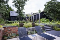 Contemporary garden in July featuring loungers on a terrace with steps leading up to an oak pergola. 
