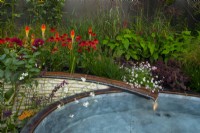 A rill flowing into a pool surrounded by autumnal plants,   Echinacea 'Eccentric', Persicaria amplexicaulia 'Fire Dance' Erigeron 'Lavender Lady, Dahlia 'Black Narcissus', Sedum 'Jose Aubergine', Kniphofia and Stipa giganta in the Finding Our Way Garden.