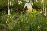 An egg shaped oak hide in woodland area surrounded by Betula pendula, and plants including Rudbeckia laciniata 'Herbstonne' - cone flower,  Calamagrositis brachytrichia - Korean feather grass and Miscanthus sinensis in the Yeo Valley Organic Garden.