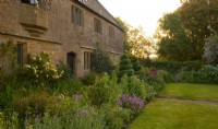 Rosa 'The Pilgrim', Spiral shaped Buxus topiary and a border of Papaver orientale, Gladiolus communis and Phuopsis stylosa in front of Lower Severalls Farmhouse at sunrise.