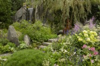 Colourful plants on a hillside beside a waterfall in 60 Degrees East, a garden between continents. Trees: Salix alba 'Tristis' and Pinus cembra - Stone Pine.  Sculptures: Penny Hardy
