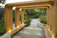 A modern timber structure with benches leading to a paved area surrounded by rills and raised herbaceous borders containing autumn planting in the Finding Our Way, An NHS Tribute Garden 