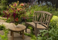 An autumnal flower arrangement of Dahlias, Persicaria, Helianthus and Acalypha Hispida - chenille plant - next to a wooden garden seat at the Gaze Burvill trade stand.