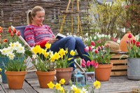 Woman relaxing on chair surrounded by containers of tulips and daffodils.