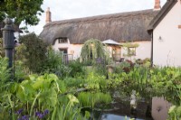 View of the garden with pond in foreground backed by thatched house