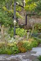 The M and G Garden. Paved area with a mix of gravel and paving for improved drainage. Featuring urban industrial decorative pipework. Autumn planting combination including Acanthus hungaricus 'White Lips', Calamgrostis brachytricha, Aralia, and Artemisia lactiflora. At the back, Actaea simplex 'Atropurpurea' with Hydrangea quercifolia.