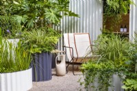 Hot Tin Roof Garden. Cream deck chair and jug with corrugated steel cream painted back wall and circular containers, including small pool. Shade-loving planting includes Fatsia japonica, Carex 'Ice Dancer', Heuchera 'Key Lime Pie', Vinca minor 'Bowles Purple', and Geranium 'Rozanne', with Pontederia lanceolata in the pond. Ferns include Polypodium vulgare and Dryopteris erythrosora 'Brilliance'.