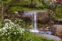 Psalm 23 Garden. Naturalistic cascade of water from granite, worn rocks in drystone wall into tranquil pool. Featuring Eupatorium 'Lucky Melody', Carex sp.,  and young shoots of Viburnum opulus .