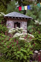 Trailfinders' 50th Anniversary Garden. Stone, timber, and slate building housing water-driven prayer wheel. Pine Pinus wallachiana behind, Persicaria amplexicaulis and Selinum wallichianum in front.