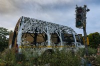 Delays Expected, by Dan Rawlings, exhibited by Saatchi Gallery. Van etched and carved into latticework of trees and plants, surrounded by planting evocative of wasteland, including white Persicaria, achillea, and brambles. Decaying traffic light in foreground.