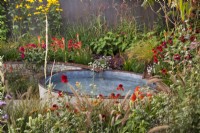 A rill flowing into a pool surrounded with colorful planting including Echinacea 'Eccentric', Persicaria amplexicaulia 'Fire Dance' Erigeron 'Lavender Lady, Dahlia 'Black Narcissus', Sedum 'Jose Aubergine', Pennisetum,  Kniphofia and Miscanthus sinensis. Finding our Way: An NHS Tribute Garden at RHS Chelsea Flower Show 2021 