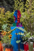 Pop Street Garden. Brightly coloured head sculpture on blue base, surrounded by plants in painted containers, including Colocasia esculenta 'Burgundy Stem, black grass Ophiopogon planiscapus 'Nigrescens', Miscanthus sinensis 'Dronning Ingrid', and Rudbeckia hirta.