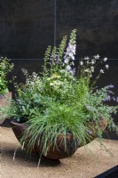 Stolen Soul Garden. Sculptural, scalloped, wooden container, with mainly green and white planting stand on a bare surface, with black wall behind, set with an amethyst crystal. Plants include white echinacea, white thalictrum, white astrantia, white Oenothera lindheimeri, delphiniums, evergreen foliage, and grasses.